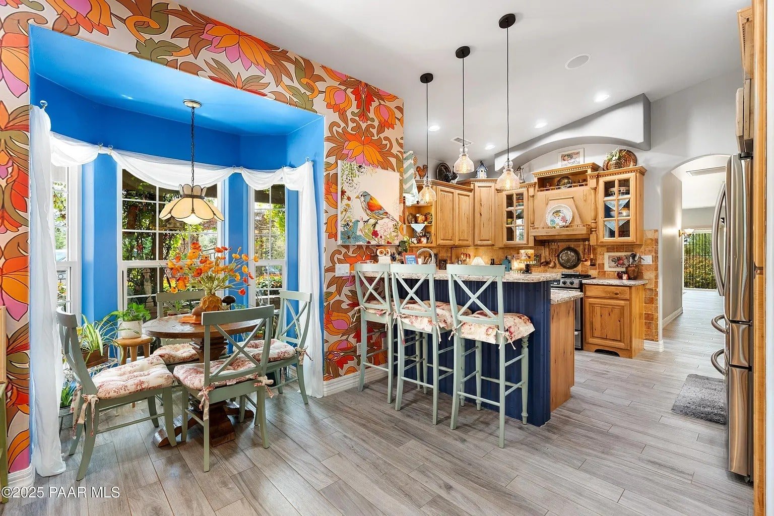 Colorful kitchen with floral wallpaper, wooden cabinets, bar stools, and a dining area by a bay window.