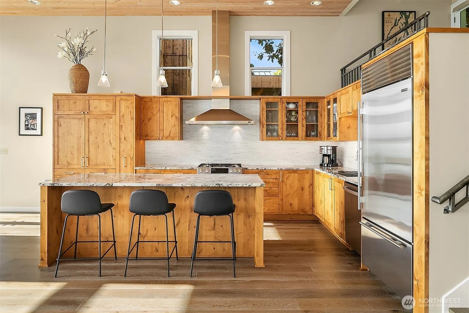 Modern kitchen with wooden cabinets, marble countertops, stainless steel appliances, and three black bar stools.