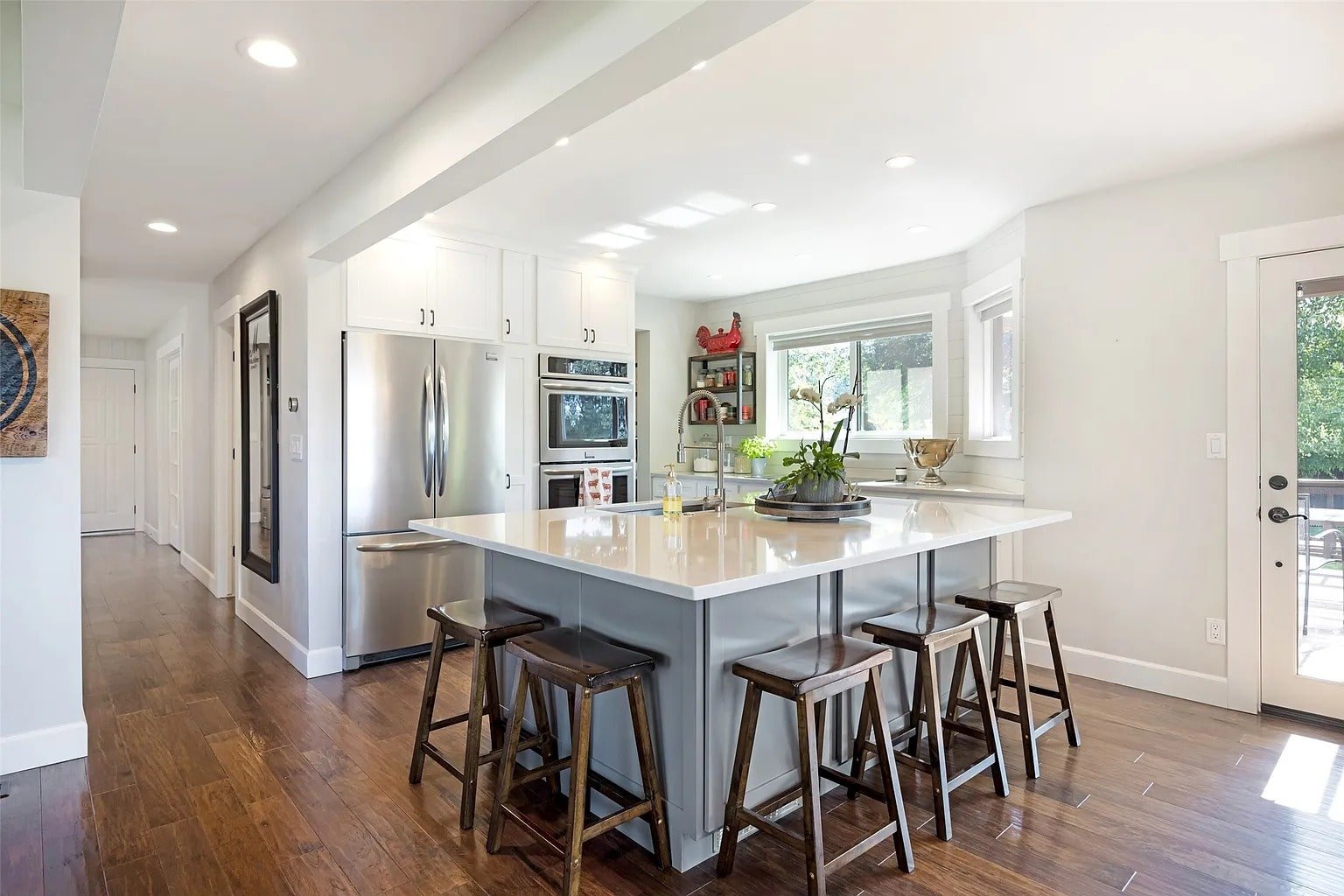 Modern kitchen with large island, wooden stools, stainless steel appliances, and bright natural lighting.