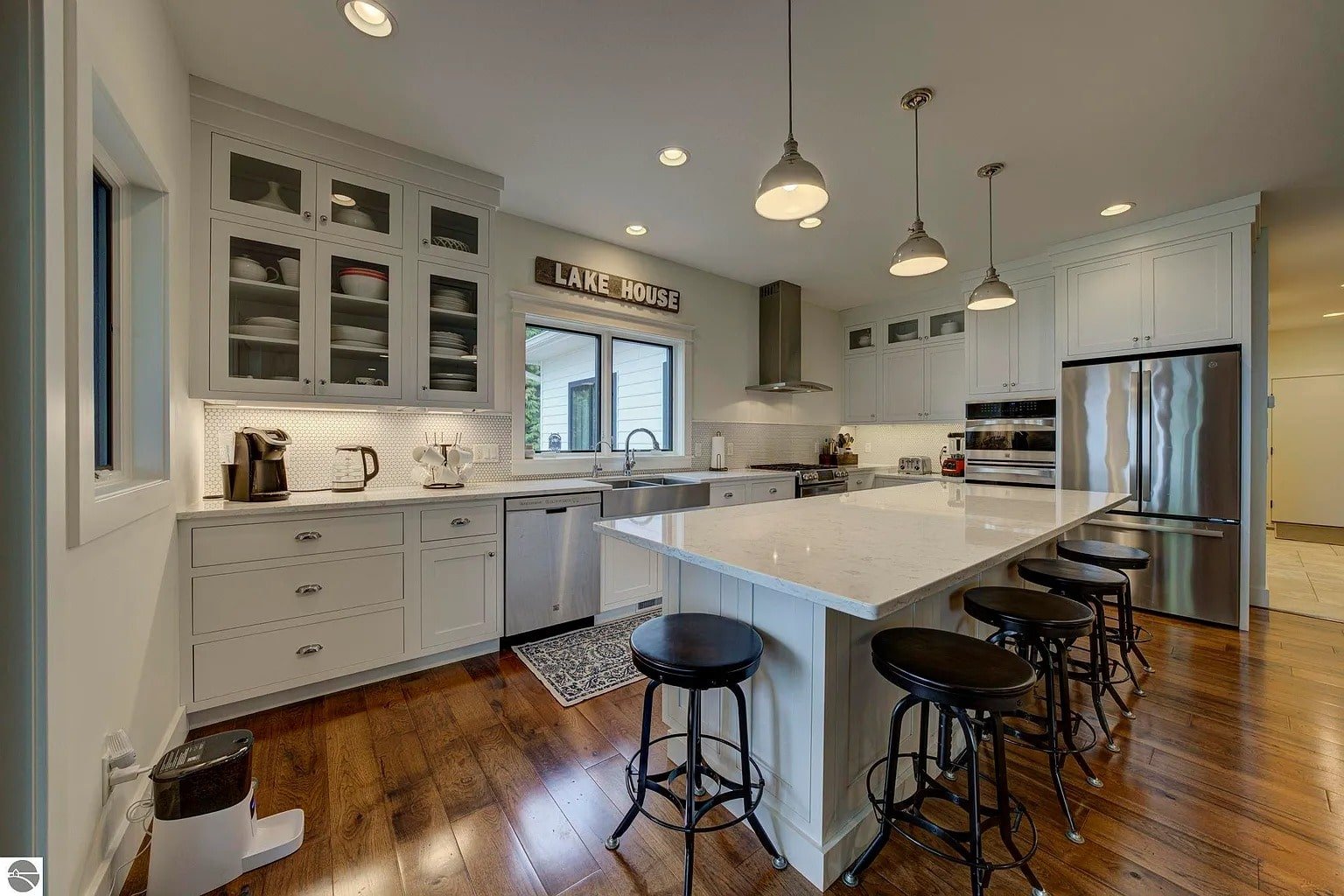 Modern kitchen with white cabinets, large island, barstools, and "Lake House" sign above the window.