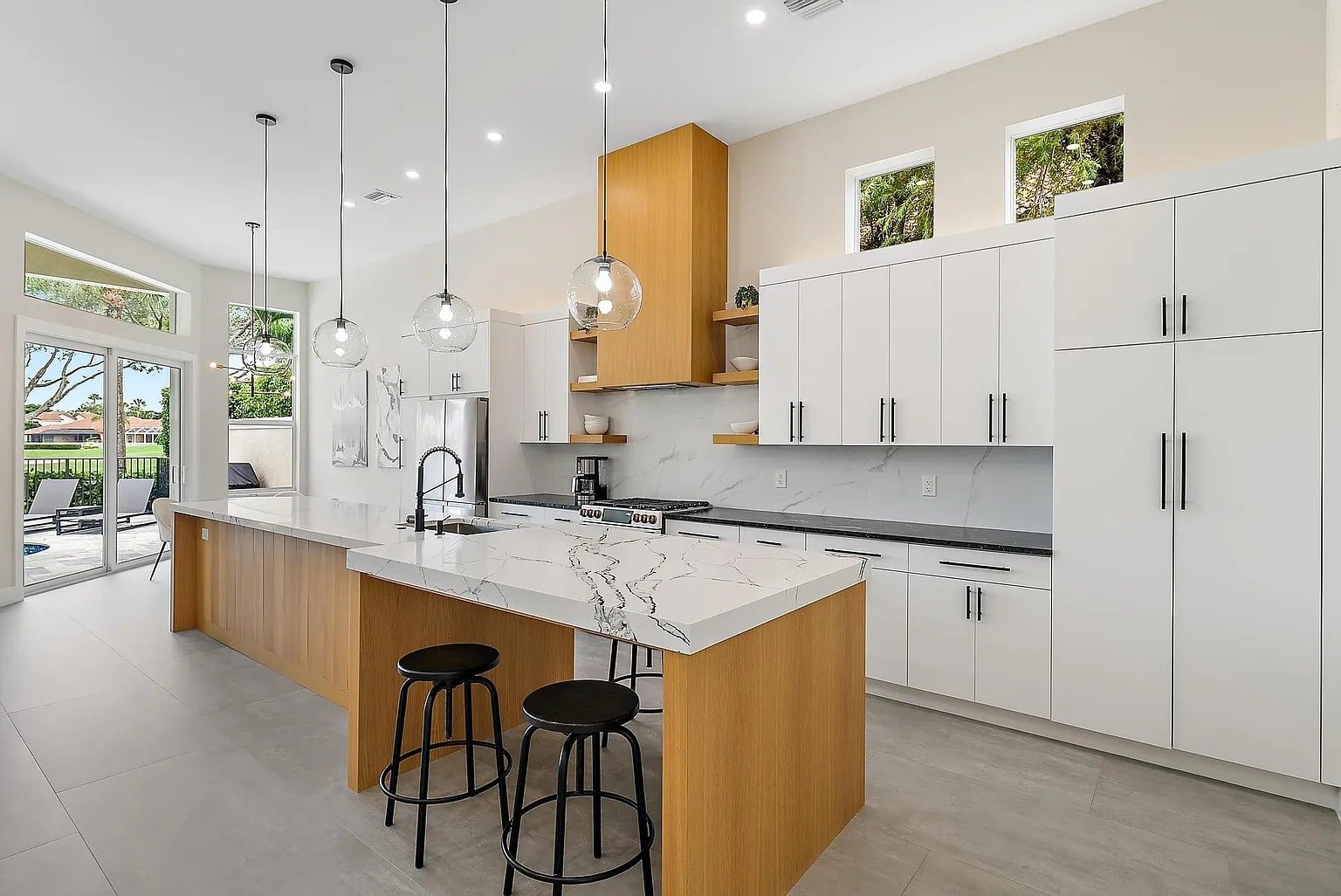 Modern kitchen with white cabinets, marble countertops, wooden accents, and pendant lights. Large windows and patio view.