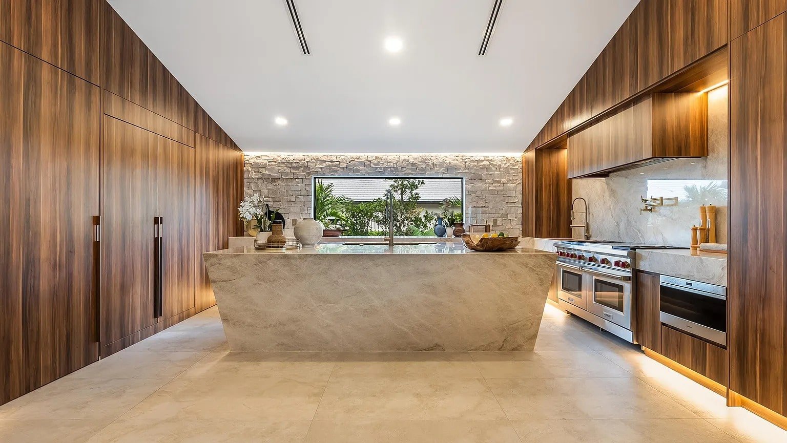 Modern kitchen with wooden cabinets, marble island, stainless steel appliances, and large window view.