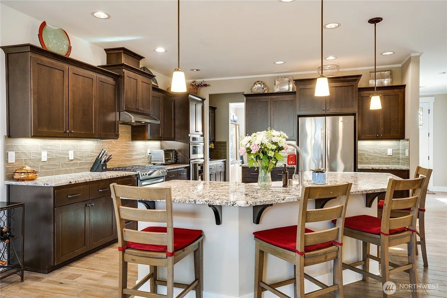 Modern kitchen with dark wood cabinets, granite countertops, stainless steel appliances, and a central island with red-cushioned chairs.