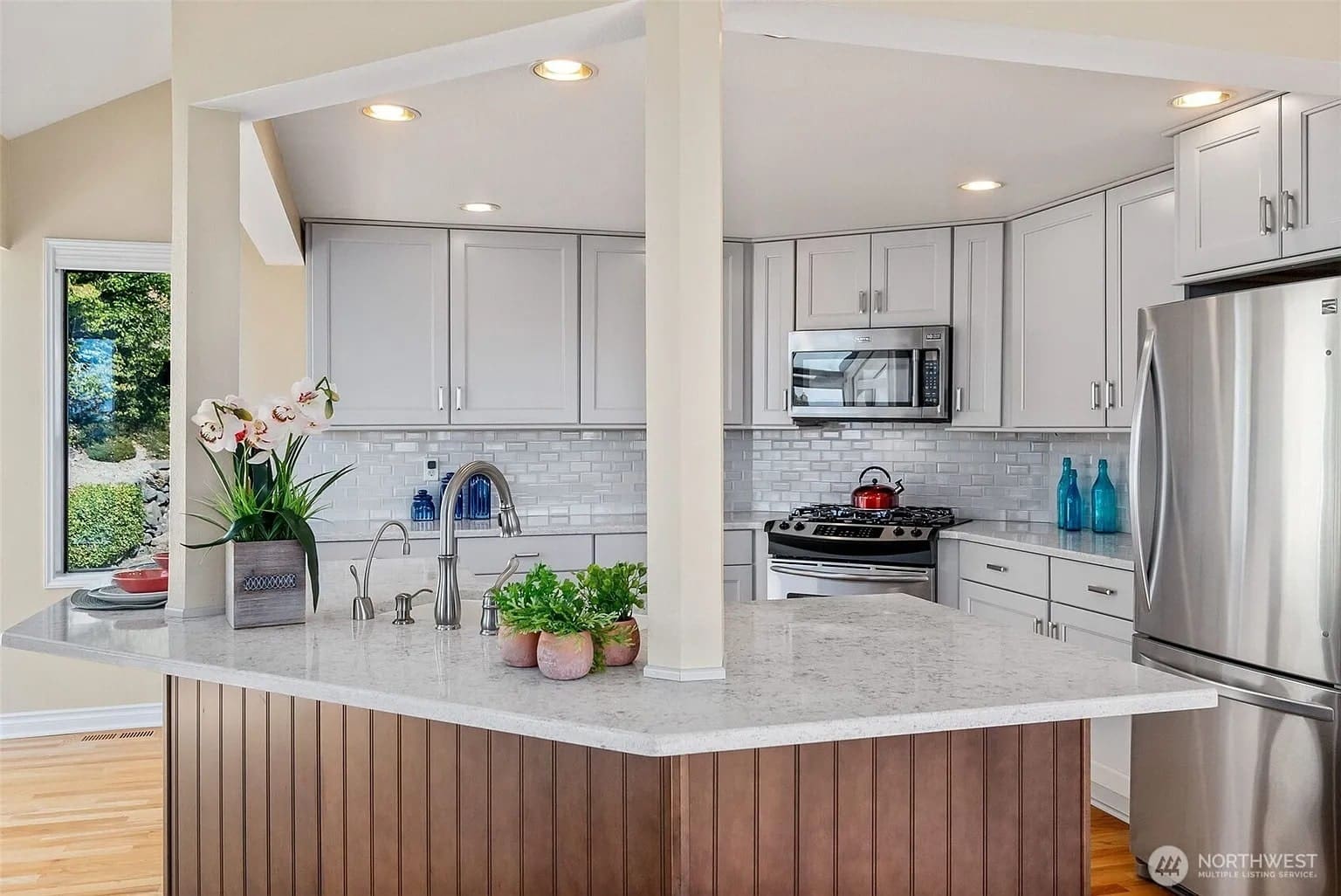 Modern kitchen with white cabinets, stainless steel appliances, and a marble countertop with plants.