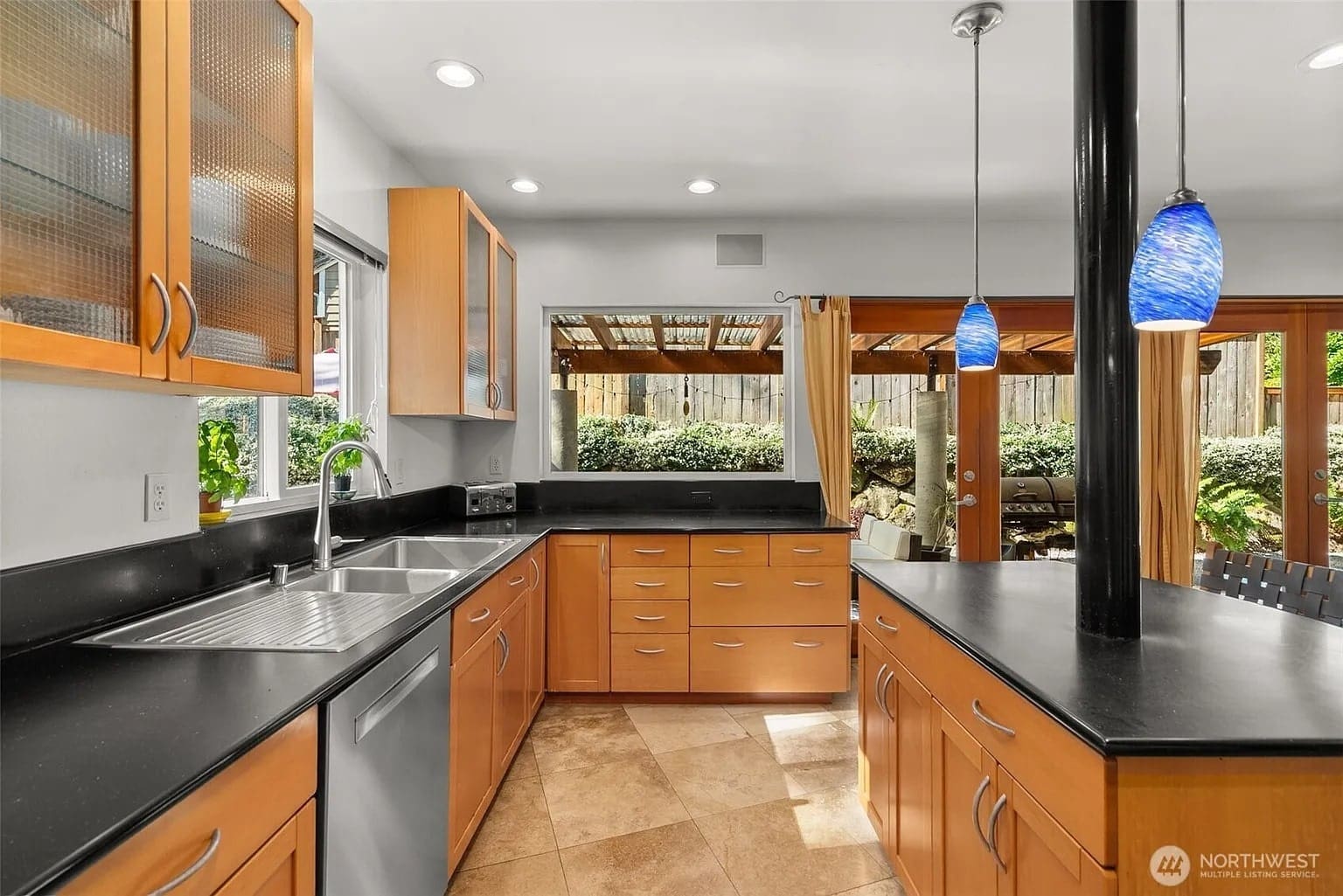Modern kitchen with wooden cabinets, black countertops, blue pendant lights, and large windows overlooking a garden.