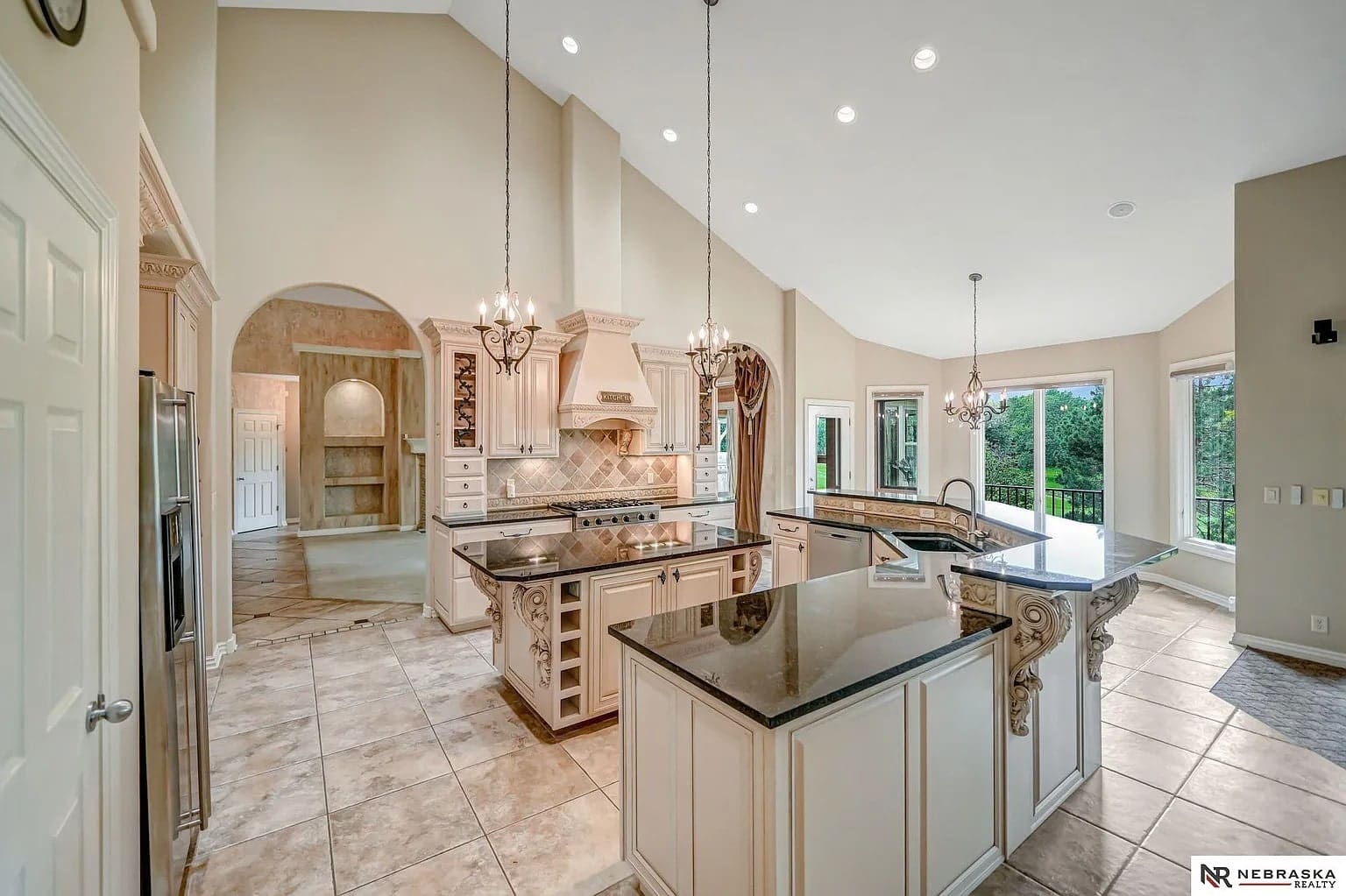 Spacious kitchen with high ceilings, marble countertops, chandeliers, and large windows overlooking greenery.