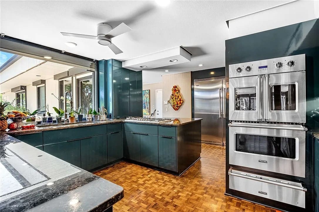 Modern kitchen with dark cabinets, stainless steel appliances, and large windows overlooking greenery.