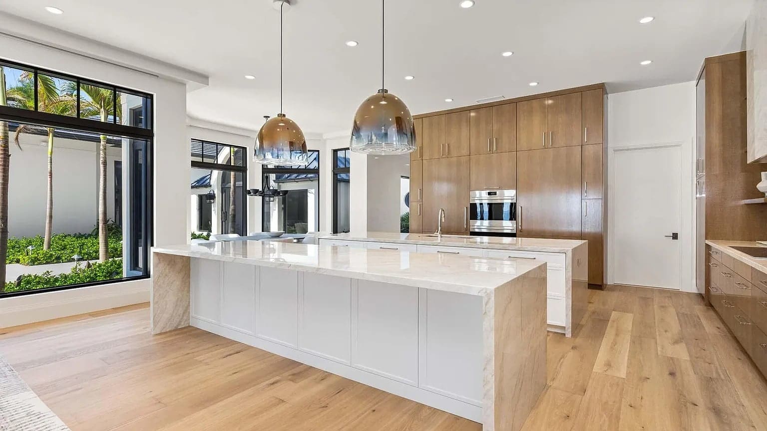 Modern kitchen with large island, pendant lights, wooden cabinets, and large windows overlooking greenery.