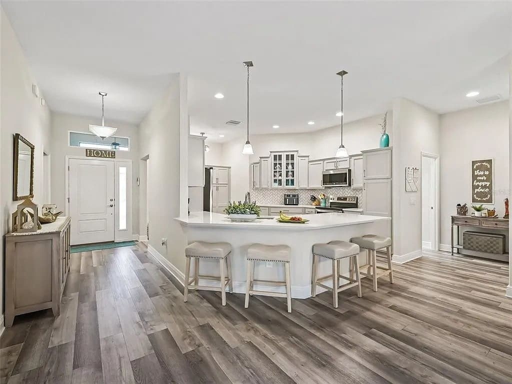 Modern kitchen with white cabinets, island with stools, and wood flooring, adjacent to entryway.