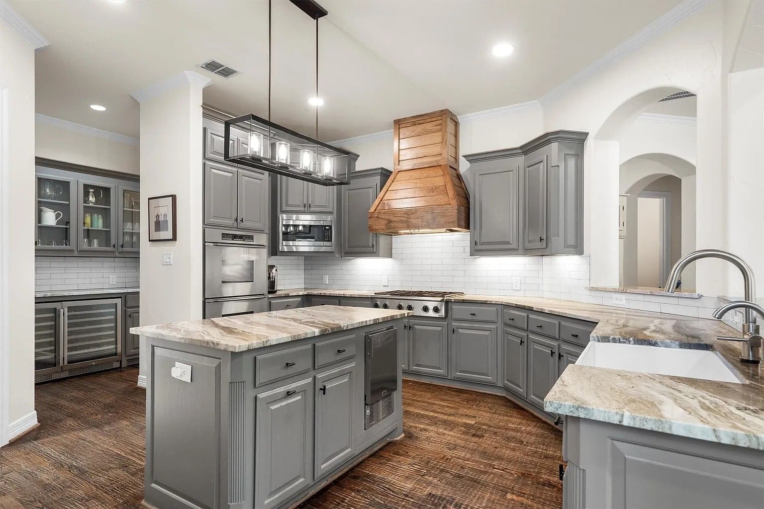 Modern kitchen with gray cabinets, marble countertops, wooden range hood, and hardwood floors.
