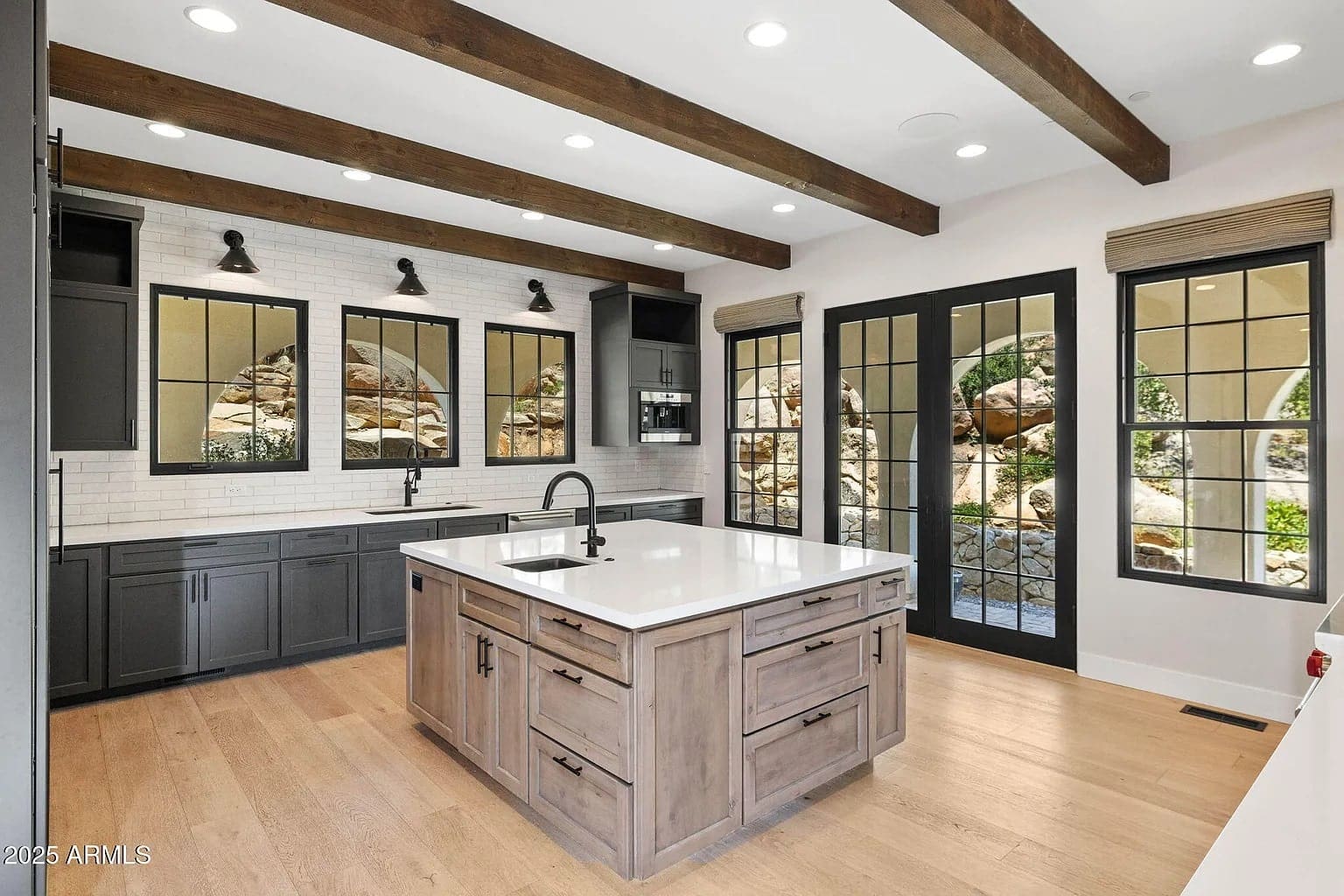 Modern kitchen with wooden beams, large island, black cabinets, and glass doors opening to a garden.
