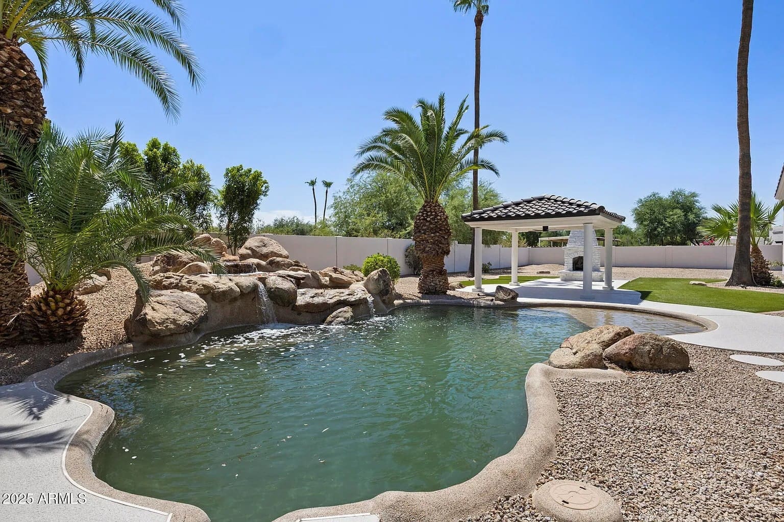 Backyard with a pool, rock waterfall, palm trees, and a small gazebo under a clear blue sky.
