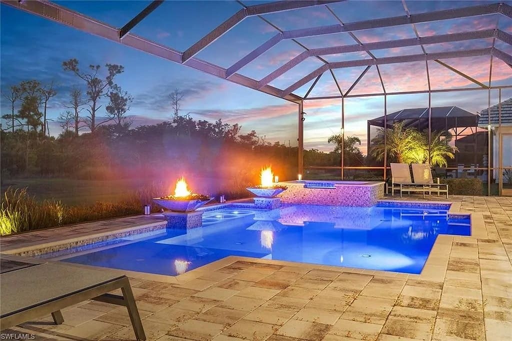 Outdoor pool area at sunset with fire features and lounge chairs under a screened enclosure.
