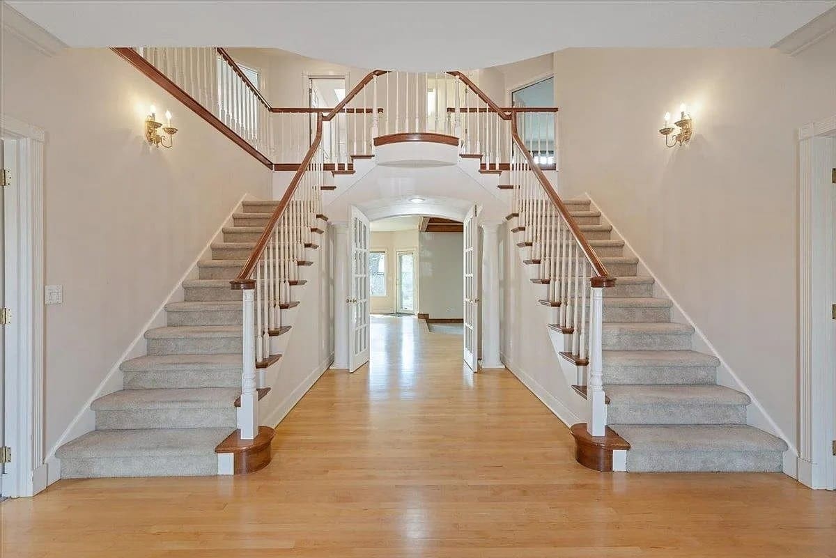 Spacious foyer with a dual staircase, wooden floors, and white walls with sconces.
