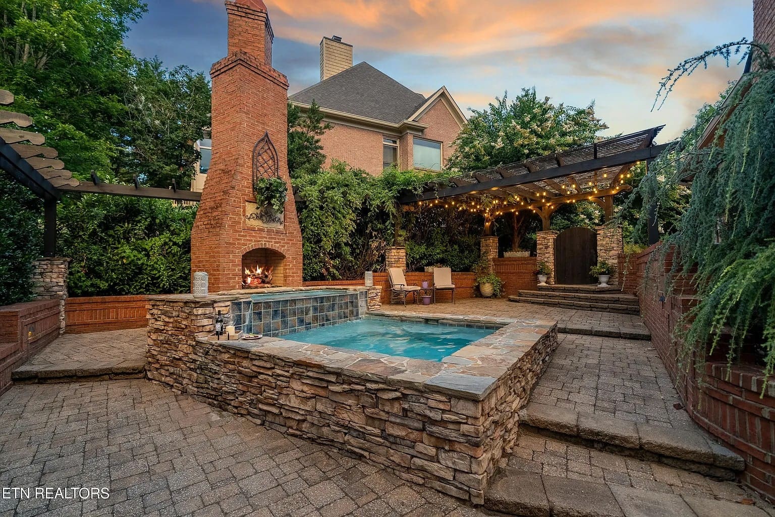 Backyard with a stone hot tub, brick fireplace, and pergola adorned with string lights.