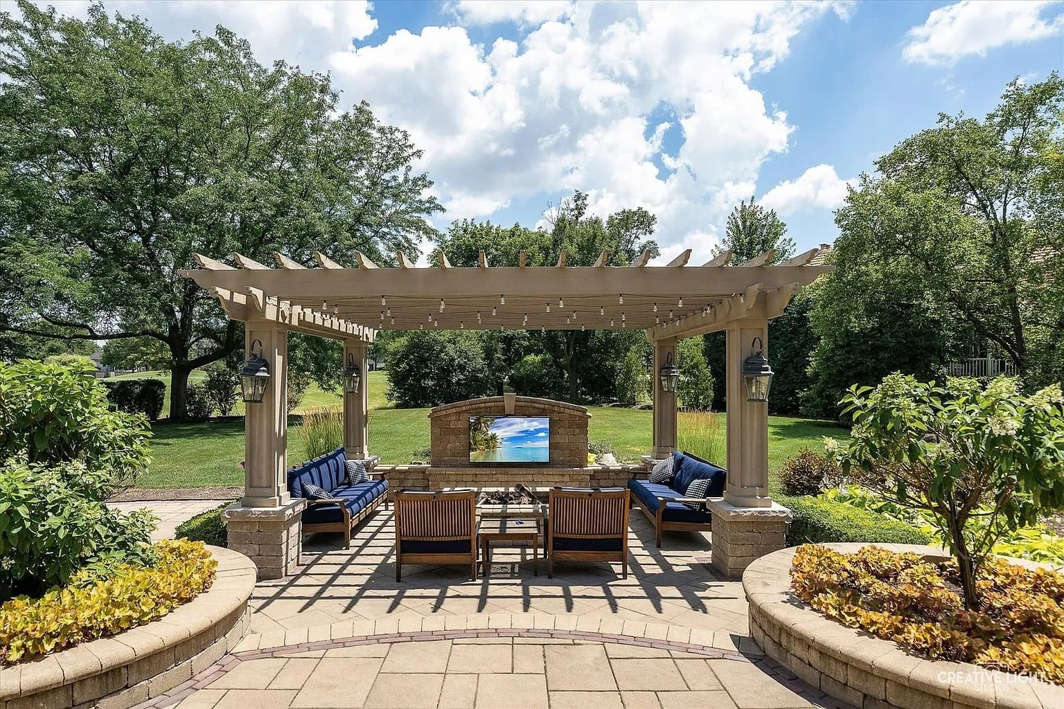 Outdoor patio with pergola, seating area, and TV, surrounded by lush greenery and trees.
