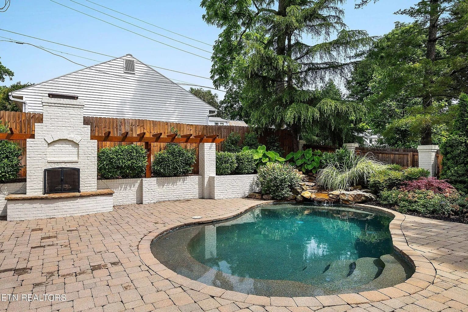 Backyard with a small pool, brick patio, fireplace, and lush greenery.