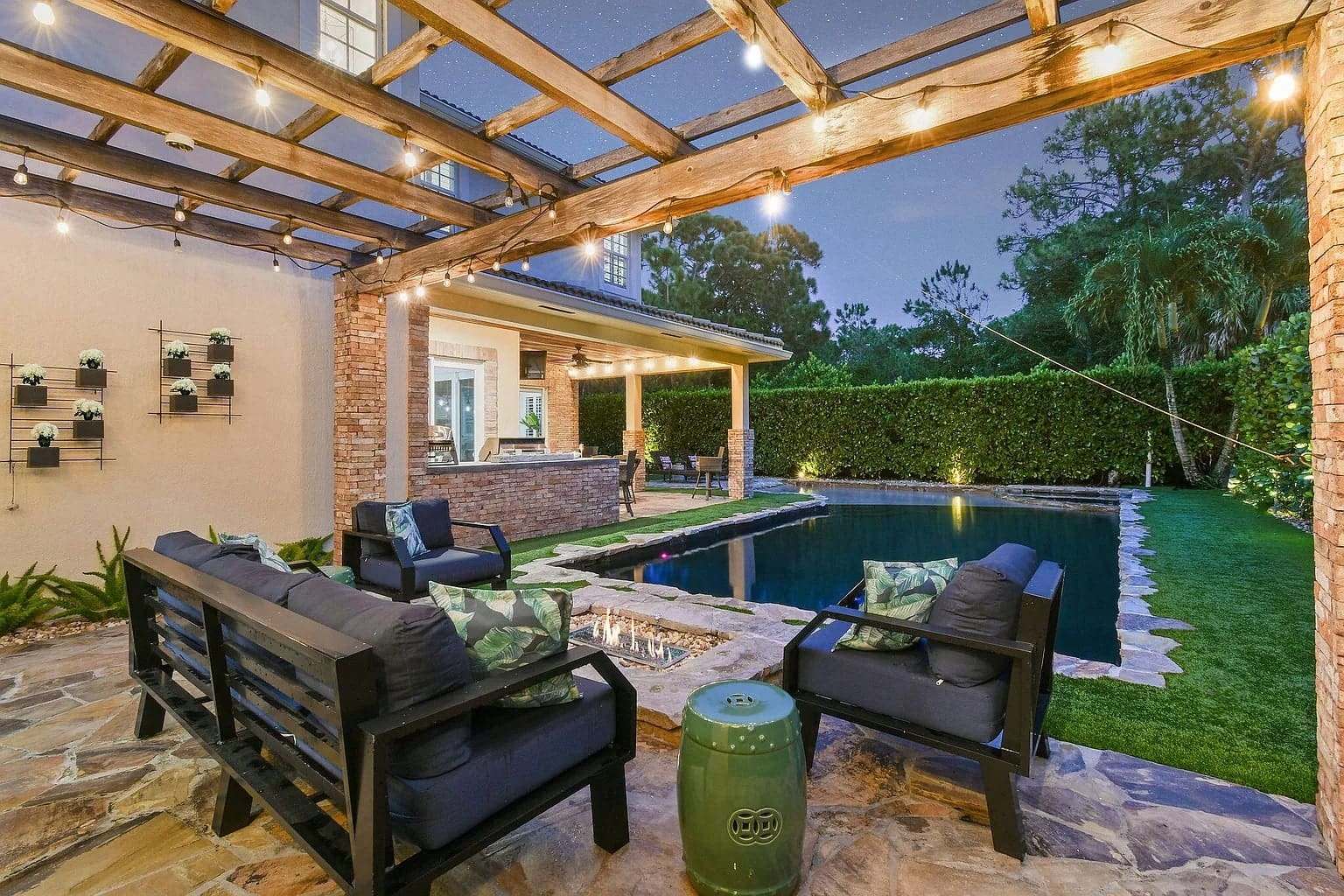 Outdoor patio with seating, pergola, string lights, and a pool surrounded by greenery at dusk.

