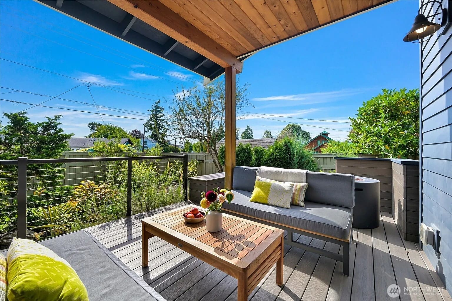 Covered outdoor patio with modern seating, wooden table, and garden view under a clear blue sky.
