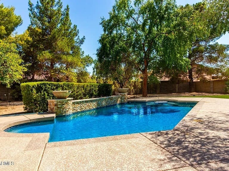 Backyard with a clear blue swimming pool, surrounded by trees and a stone wall.

