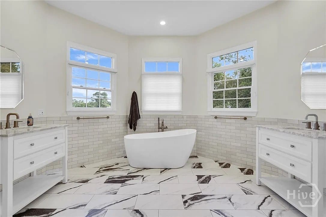 Spacious bathroom with freestanding bathtub, double vanities, large windows, and marble-patterned floor.
