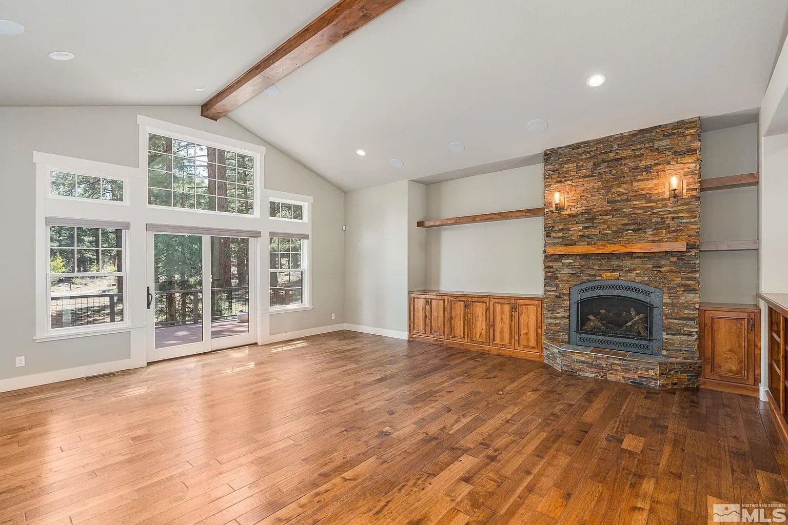 Spacious living room with wooden floors, stone fireplace, large windows, and exposed ceiling beams.