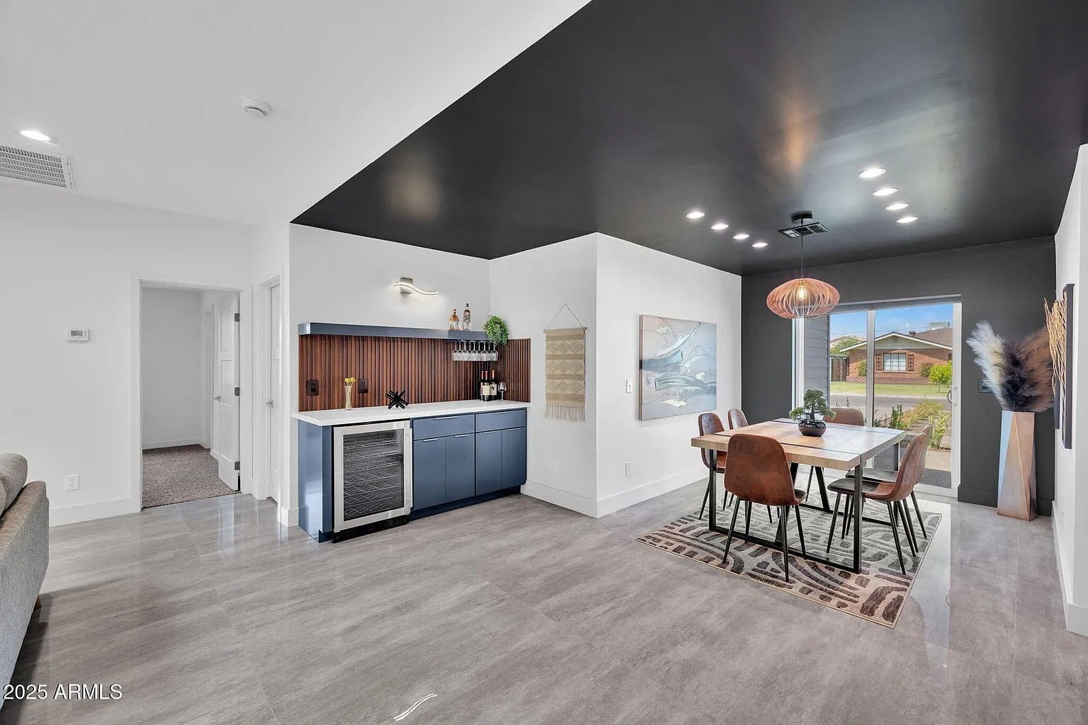 Modern dining room with dark ceiling, wooden table, chairs, and a small bar area with wine cooler.

