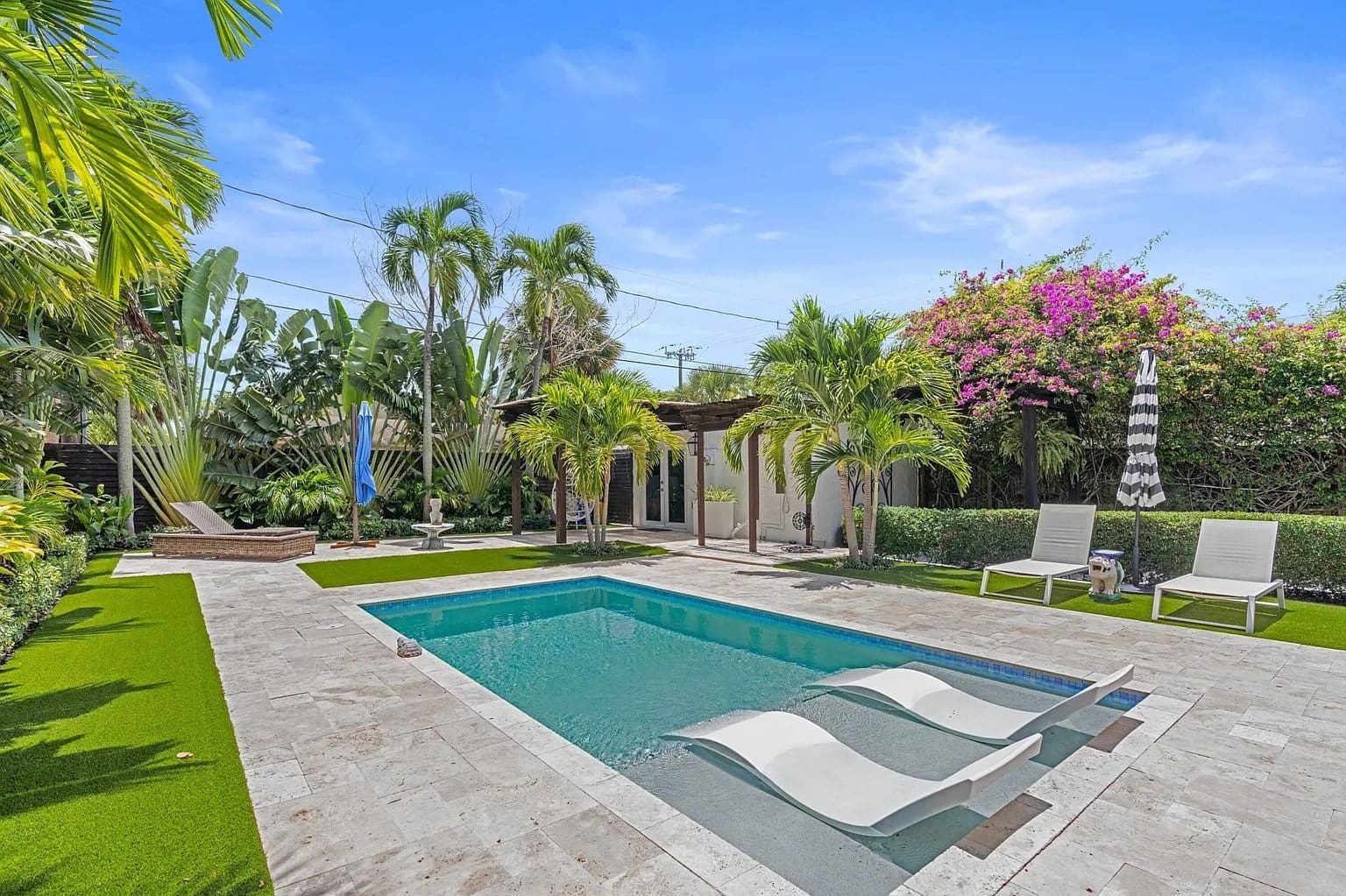 Backyard with a swimming pool, lounge chairs, palm trees, and vibrant flowering plants under a clear sky.
