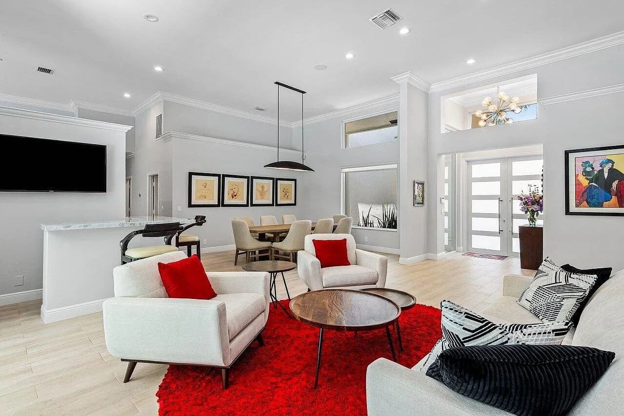 Modern living room with white chairs, red accents, dining area, and wall art. Bright and spacious.
