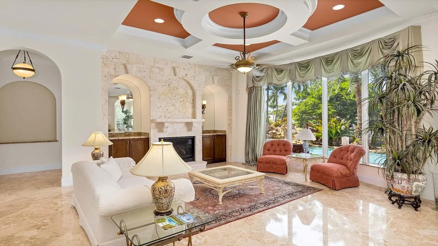 Elegant living room with large windows, plush seating, ornate ceiling, and a view of greenery outside.
