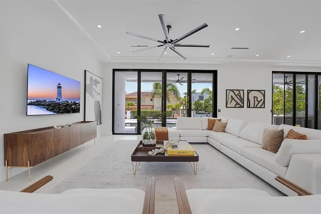 Modern living room with white sofas, large TV, ceiling fan, and glass doors opening to a patio.

