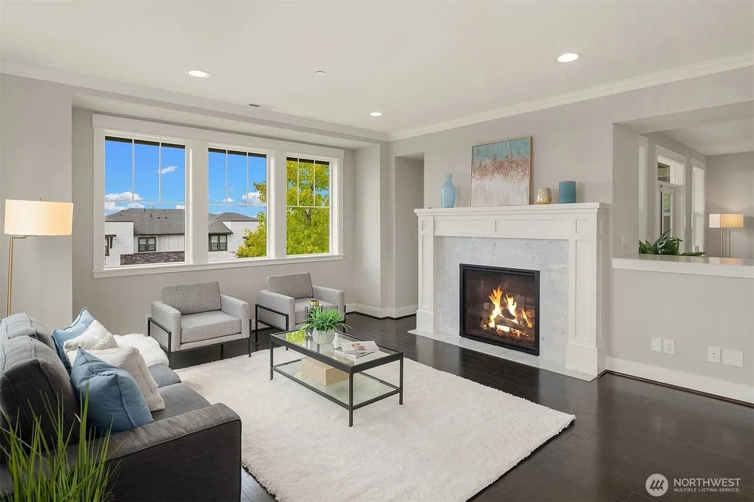 Modern living room with fireplace, gray furniture, large windows, and a white rug on dark wood floor.
