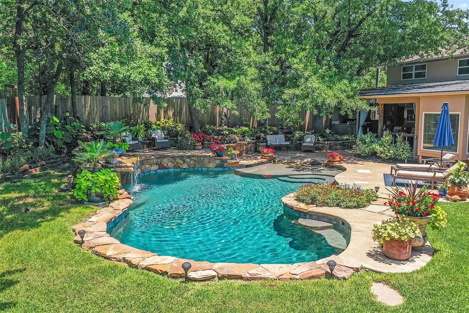 Backyard with a kidney-shaped pool, surrounded by lush greenery, flowers, and patio seating.
