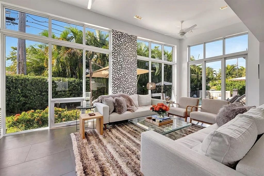 Modern living room with large windows, white furniture, patterned rug, and view of lush greenery outside.
