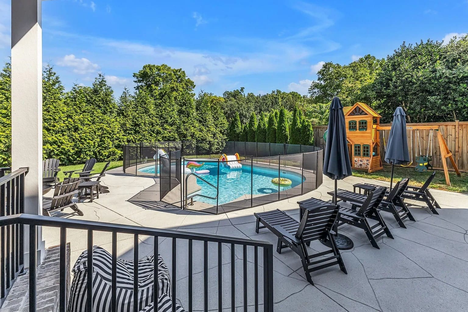 Backyard with a fenced pool, lounge chairs, umbrellas, and a wooden playhouse.
