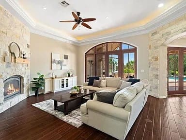 Spacious living room with stone fireplace, large windows, and a ceiling fan. Cozy seating area.
