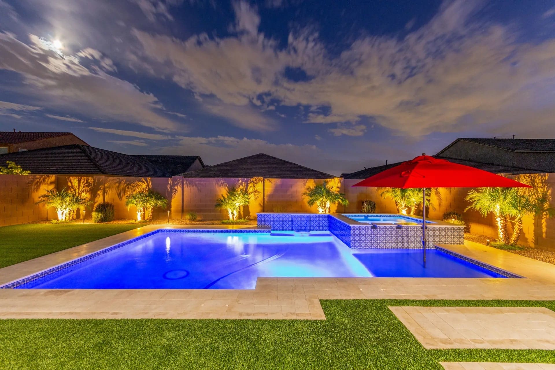 Illuminated backyard pool at night with a red umbrella, surrounded by palm trees and grass.