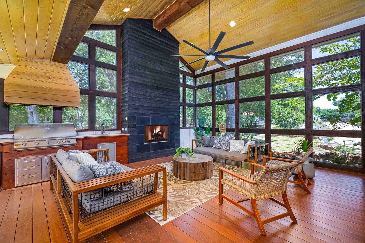 Spacious sunroom with wooden ceiling, modern furniture, fireplace, grill, and large windows overlooking greenery.
