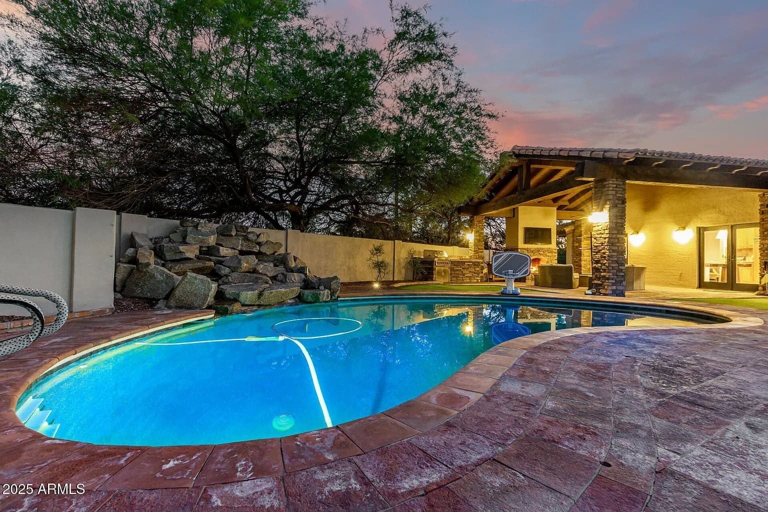 Backyard with illuminated swimming pool, rock waterfall, and a covered patio at sunset.
