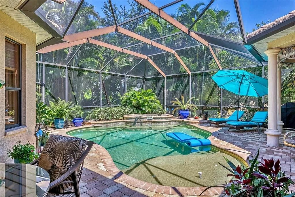Screened-in patio with a swimming pool, lounge chairs, and tropical plants under a clear blue sky.
