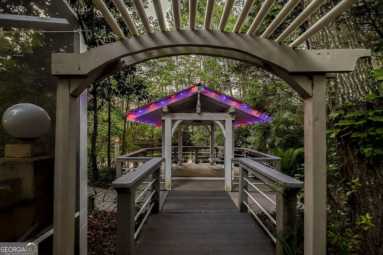 Wooden walkway with a pergola leads to a gazebo, adorned with colorful string lights.
