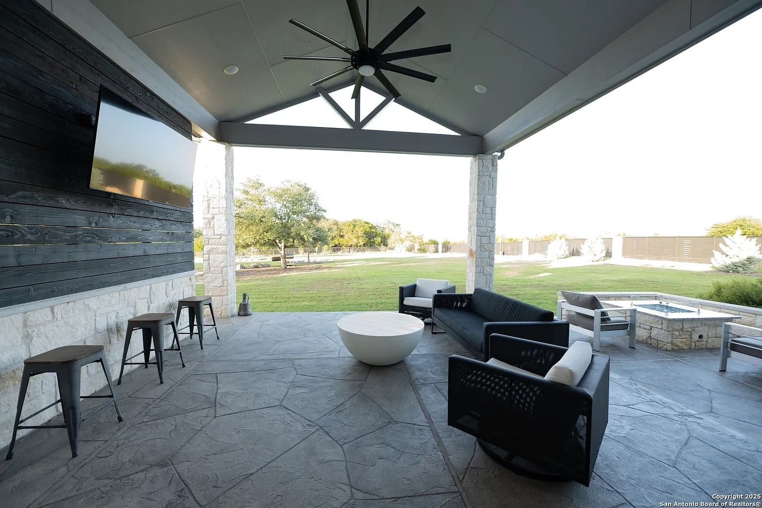 Covered patio with seating, ceiling fan, wall-mounted TV, and view of a grassy backyard.