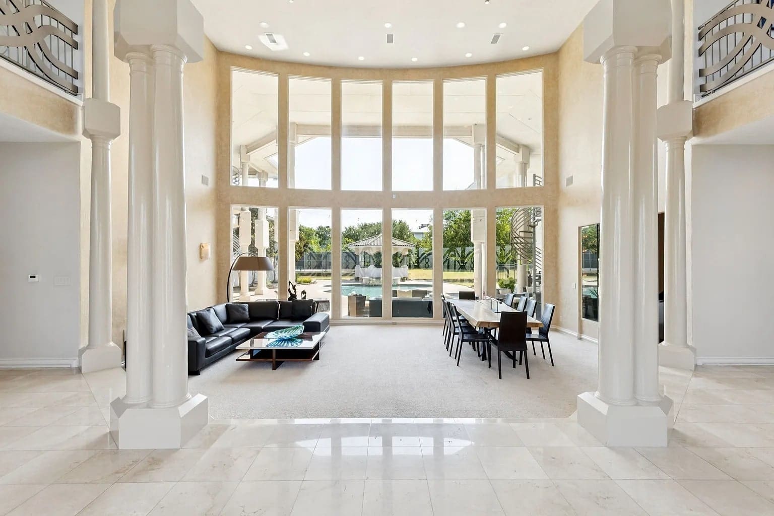 Spacious living room with large windows, black sofa, dining table, and view of a pool outside.

