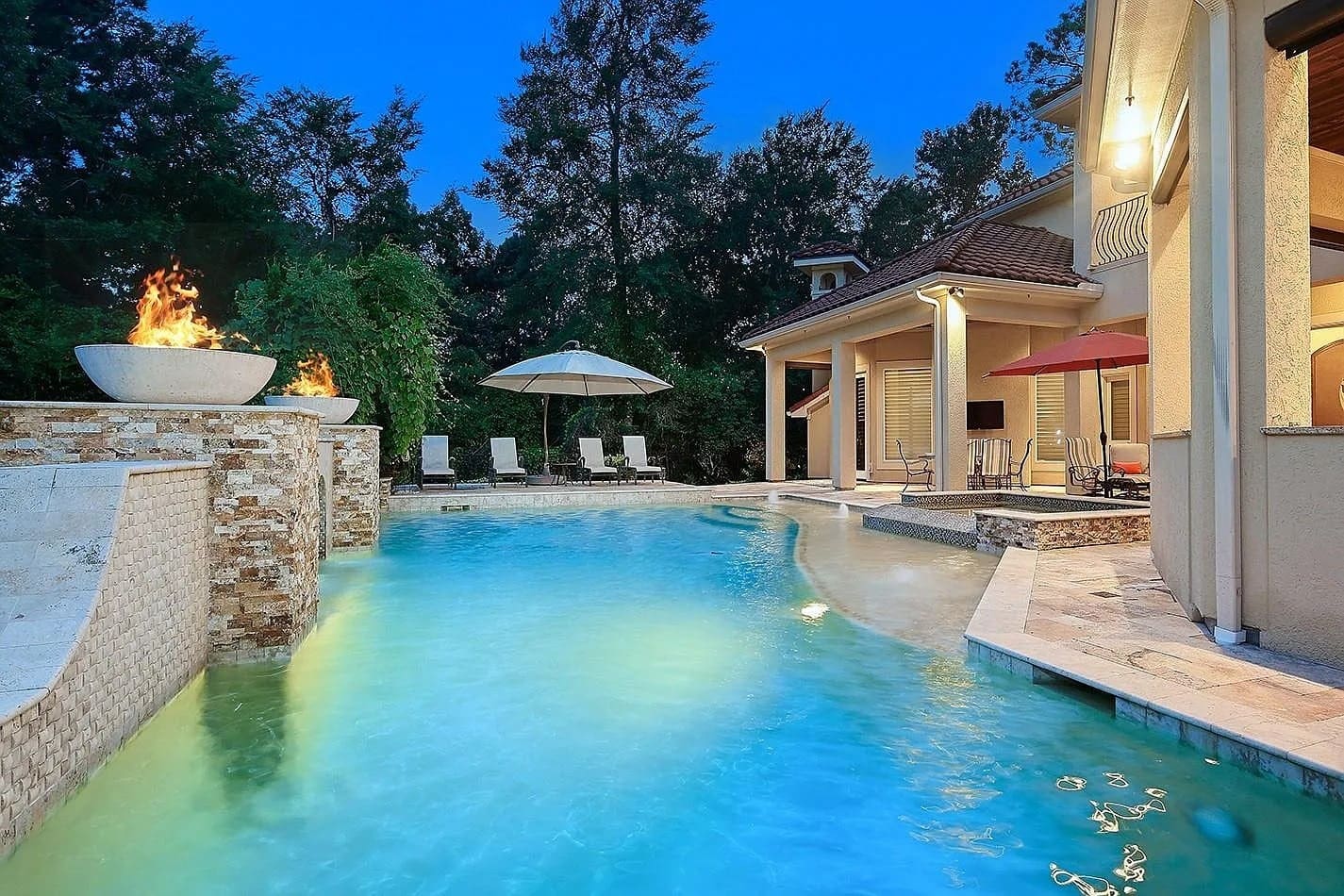 Luxurious backyard pool with waterfall, fire feature, lounge chairs, and umbrellas at dusk.
