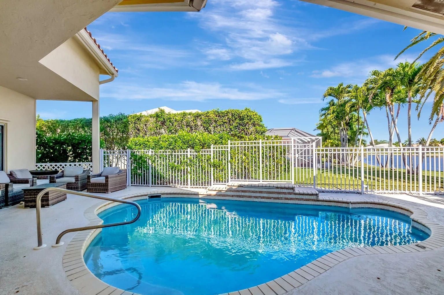 Outdoor swimming pool with safety fence, surrounded by patio furniture, palm trees, and clear blue sky.