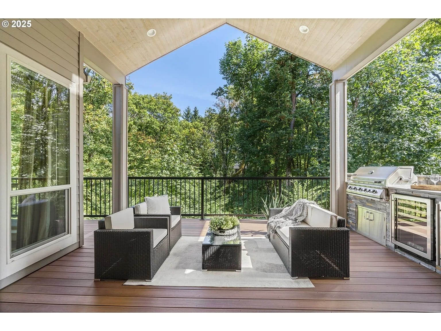 Covered outdoor patio with seating, a rug, and a grill, overlooking lush green trees.
