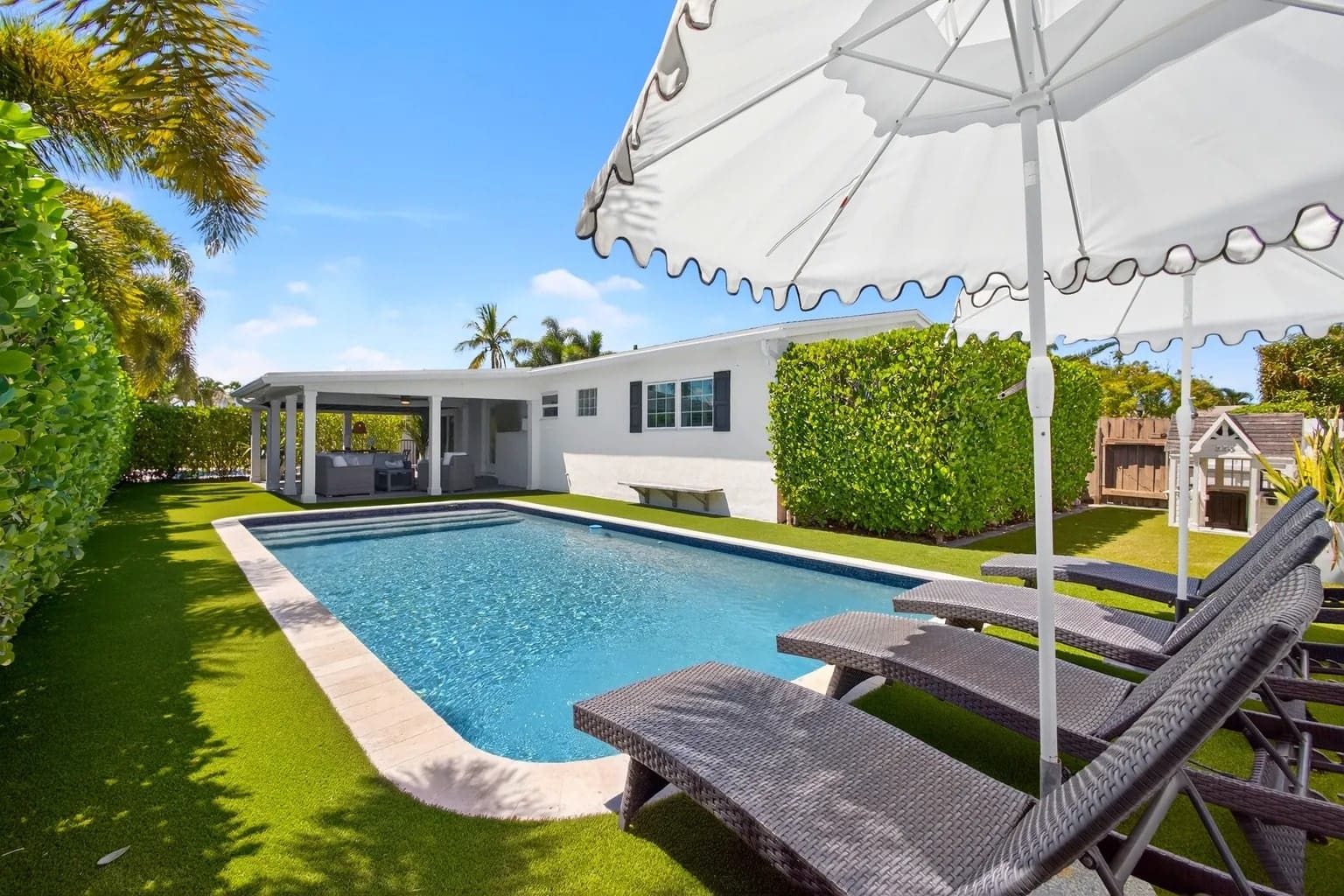 Backyard with a swimming pool, lounge chairs, umbrella, and a small playhouse, surrounded by greenery.
