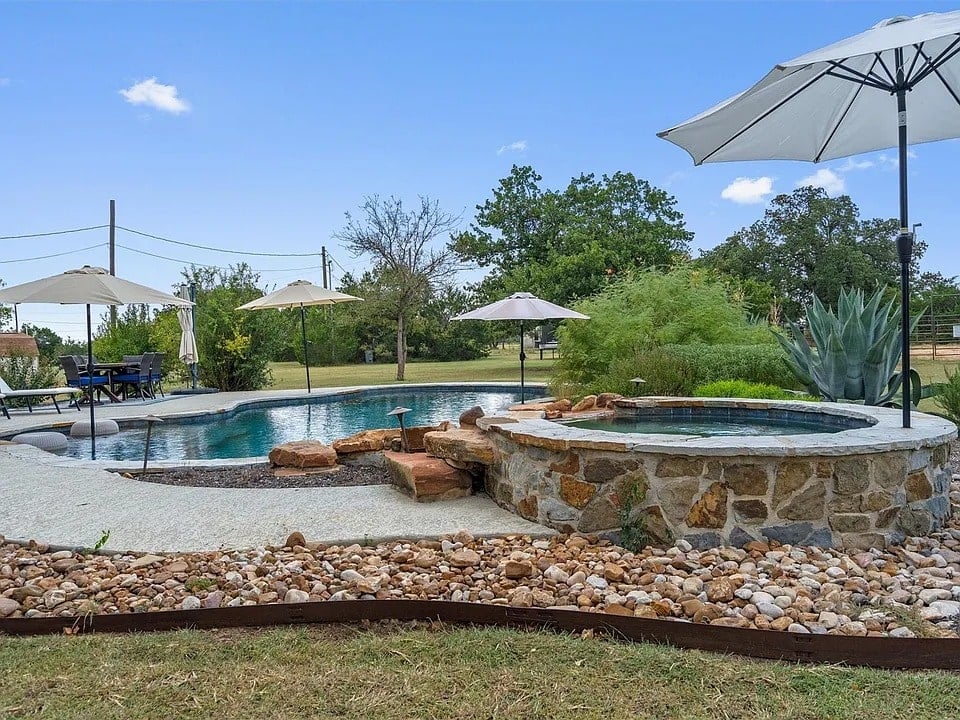 Backyard pool with stone hot tub, surrounded by umbrellas and greenery under a clear sky.