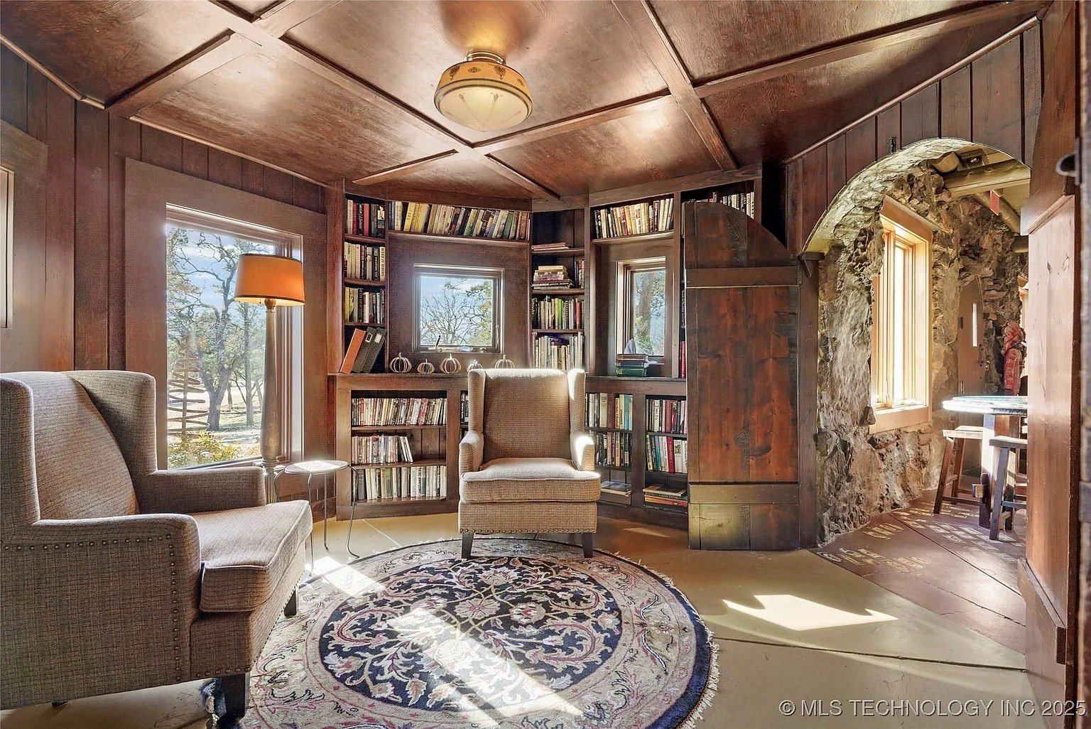Cozy library with armchairs, bookshelves, large windows, and a round rug on wooden floor.