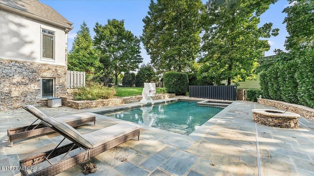 Backyard with a stone patio, pool, lounge chairs, and surrounding trees.